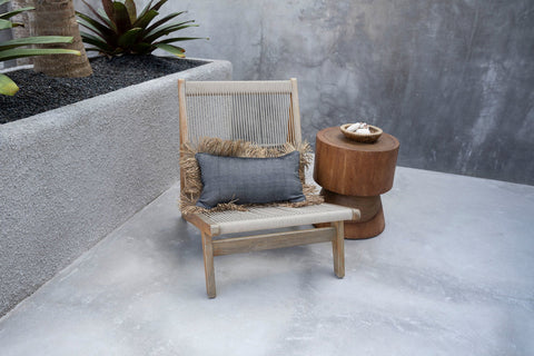 Het kussen ligt op een houten stoel op een overdekt grijs terras. Rondom staan terracotta potten met olijfbomen en natuurlijke accessoires, waardoor een mediterrane en ontspannen sfeer ontstaat.