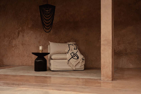 Bazar Bizar The Evil Eye plaid over een beige fauteuil naast een bruin houten bijzettafeltje.