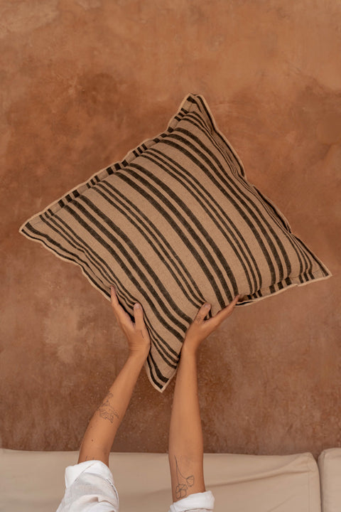 Bazar Bizar The Nordic Stripe kussen omhoog gehouden tegen een warme beige achtergrond.
