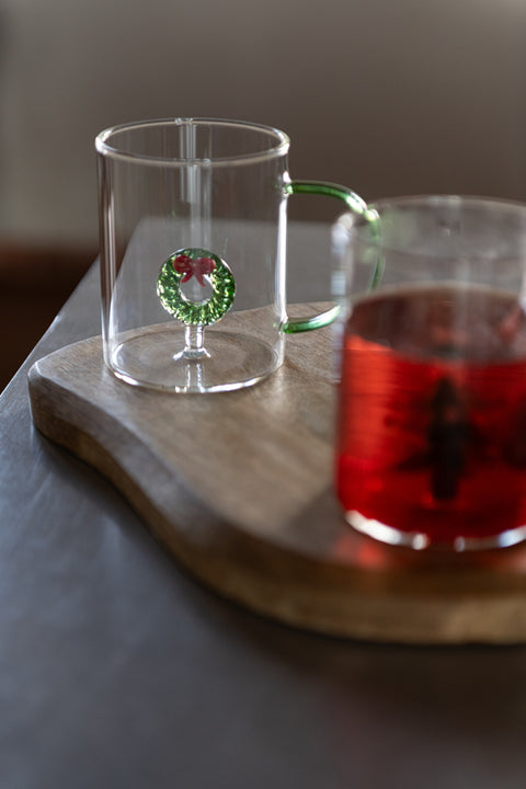 J-Line glas met kerstkrans-figuurtje op houten plankje naast een glas gevuld met thee, sfeervolle kerstsetting.