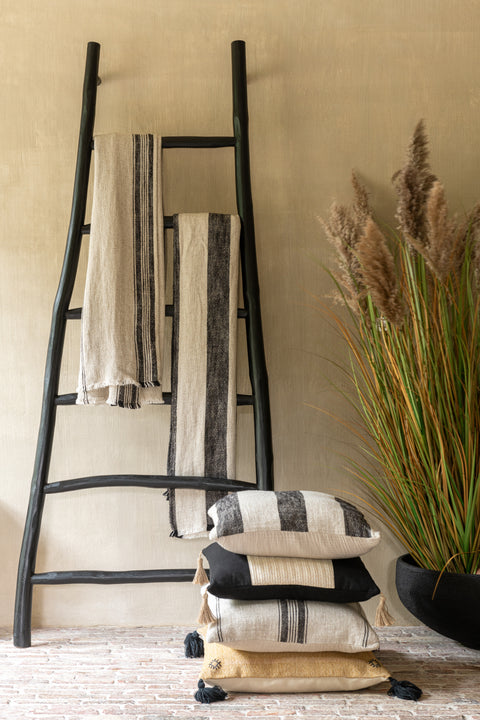 Sfeervolle interieurfoto van het J-Line Kussen ‘Linen Beige’ op de grond samen met andere kussens, tegen een houten ladder met twee plaids.