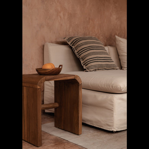 The Arc bijzettafel van naturel teakhout naast een beige zetel met gestreept kussen